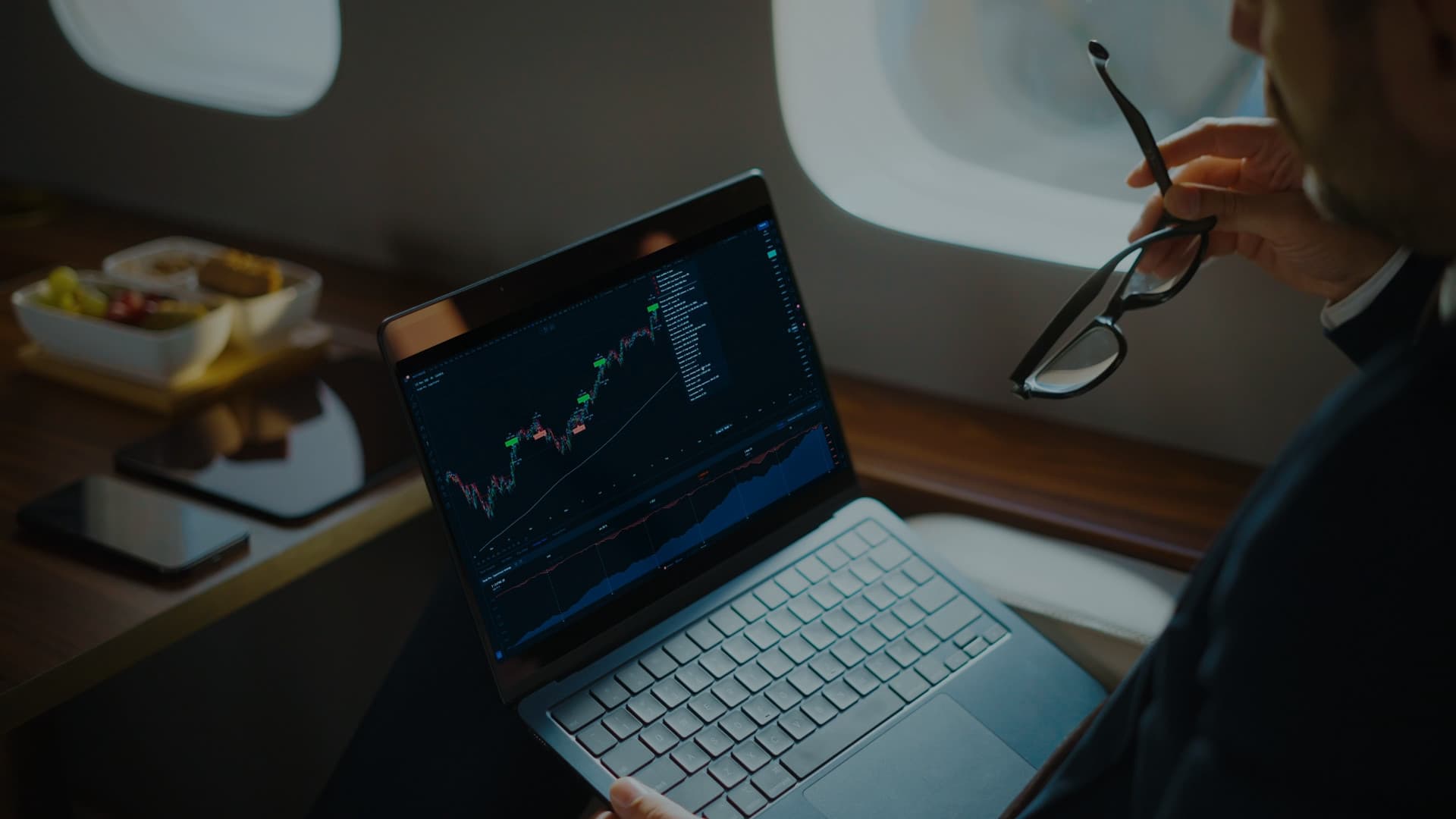 A traveller in a suit watching a candlestick chart on a laptop from an aircraft seat, with the TrackaTrader wordmark over it